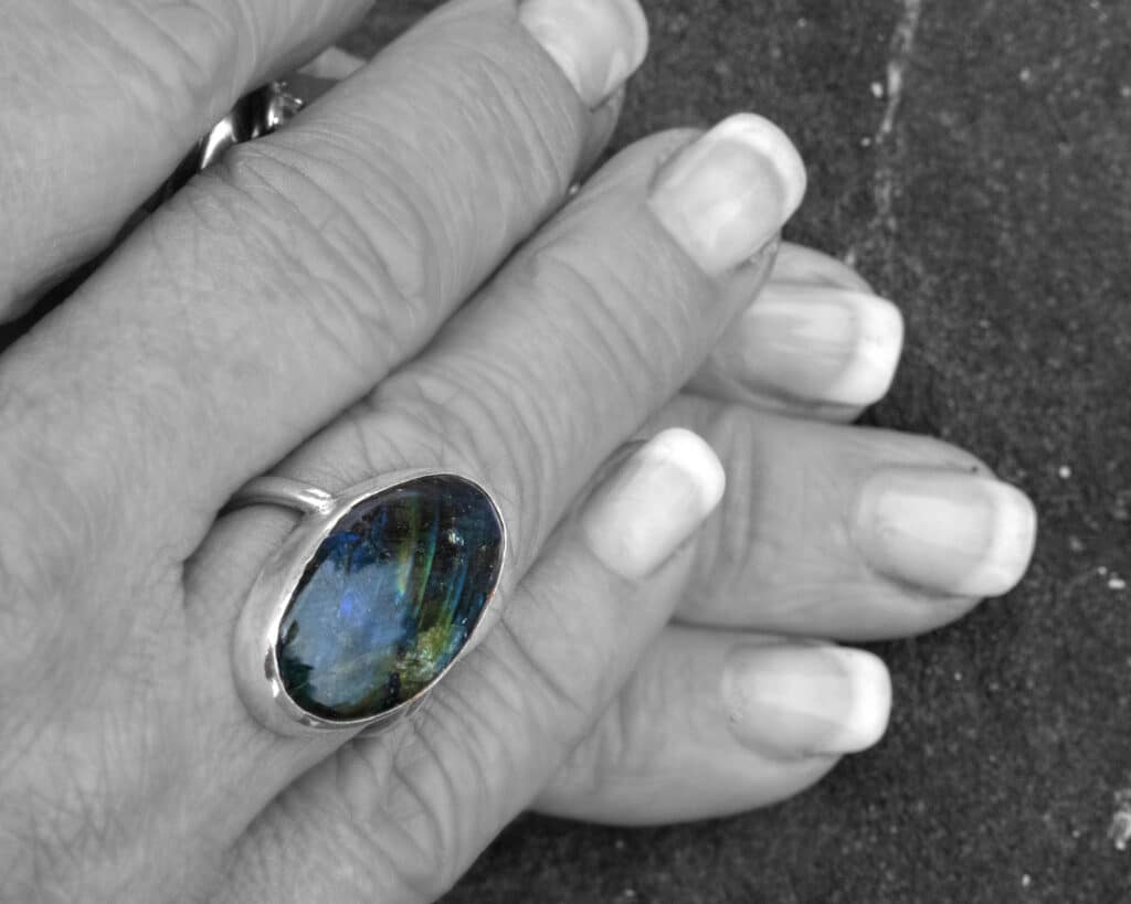 A labradorite ring with a shimmering blue-green sheen, worn on a pair of hands with natural lighting highlighting the stone’s unique flashes of color.
