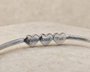 Silver memorial bangle engraved with the names Mum, Dad and Pat on three heart charms.
